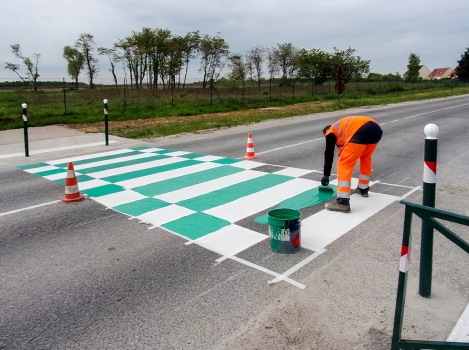 Technicien en gilet haute visibilité orange **VIA ROUTE** applique un marquage routier au sol sur chaussée neuve en enrobé bitumineux noir, à l'aide d'une **machine de marquage thermoplastique** tractée