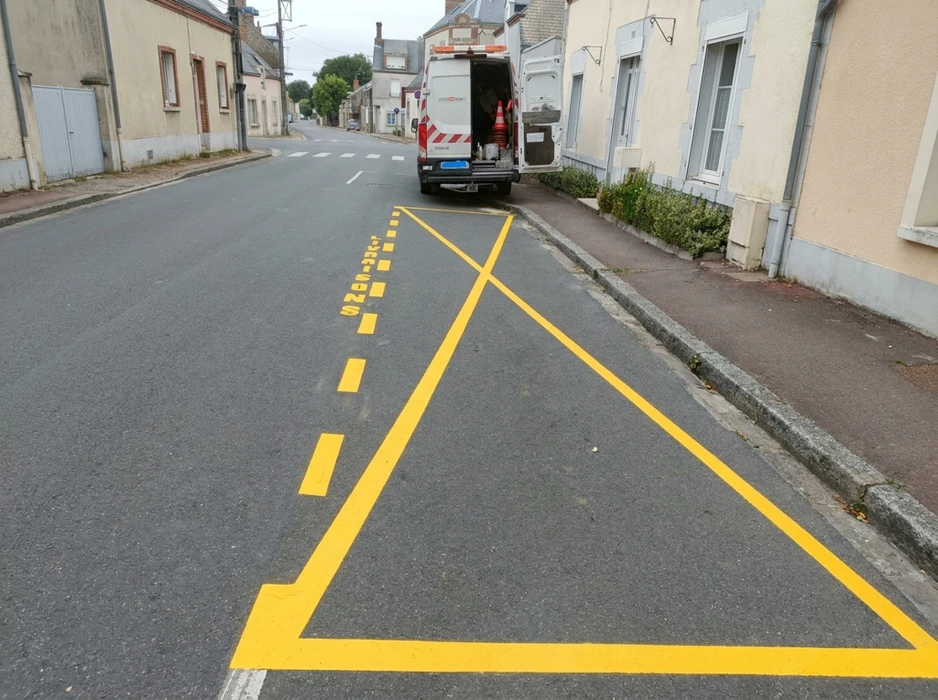Technicien en gilet haute visibilité orange marqué "VIA ROUTE" applique un marquage au sol sur chaussée neuve en enrobé bitumineux noir, à l'aide d'une machine de thermoglacage tractée