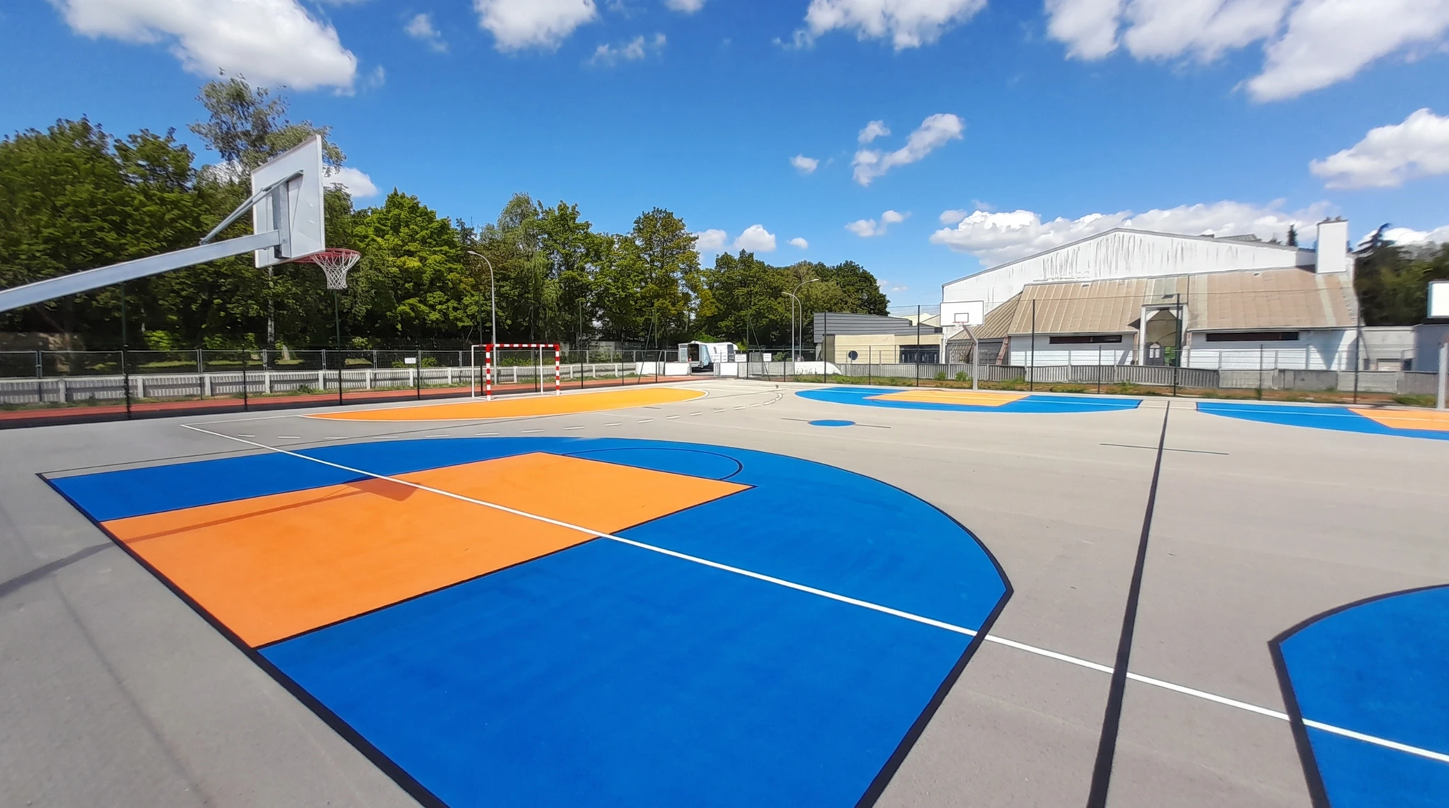 Photo aérienne ou vue en plongée d'un terrain multisports extérieur avec marquage au sol réglementaire blanc et coloré en peinture thermoplastique. Le terrain doit montrer clairement les délimitations nettes d'au moins deux sports distincts (terrain de basket-ball avec ses lignes blanches caractéristiques, terrain de handball ou de volley-ball avec filet). Revêtement en enrobé bitumineux ou béton couleur. Bordures claires, environnement urbain ou scolaire. Aucun enfant. Finition professionnelle moderne, bonne définition, jour dégagé.
