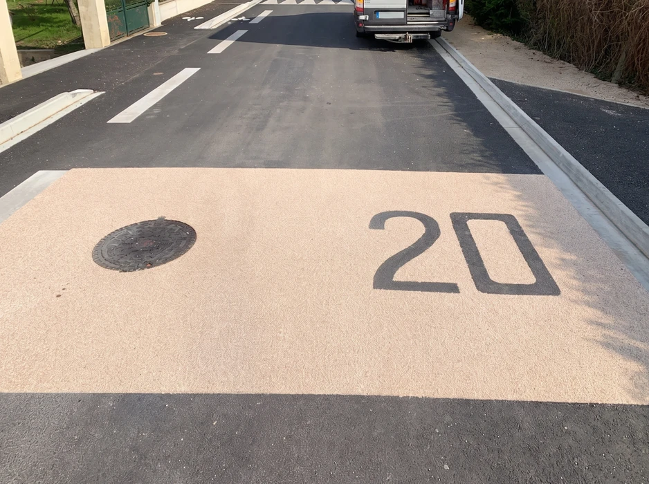 Technicien en gilet haute visibilité orange marqué "VIA ROUTE" applique un marquage au sol sur chaussée neuve en enrobé bitumineux noir, à l'aide d'une machine de thermoglacage tractée