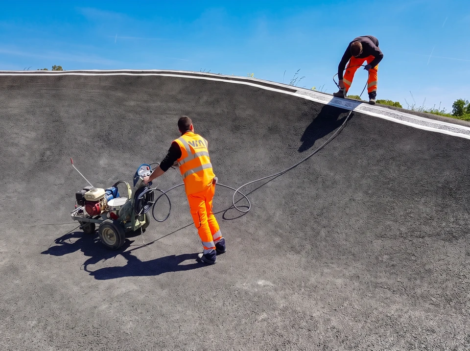 Deux techniciens en gilets haute visibilité orange marqué "VIA ROUTE", travaillant en équipe sur chantier en extérieur. Le premier applique un marquage au sol blanc thermoplastique sur asphalte noir neuf à l'aide d'une machine de traçage. Le second, à proximité, positionne des balises de sécurité ou des repères. Environnement urbain diurne, conditions ensoleillées. Les deux collaborateurs interagissent positivement, illustrant la complémentarité et le travail d'équipe. Chaussée, mobilier urbain et signalisation routière visible en arrière-plan. Photographie réaliste de qualité professionnelle.